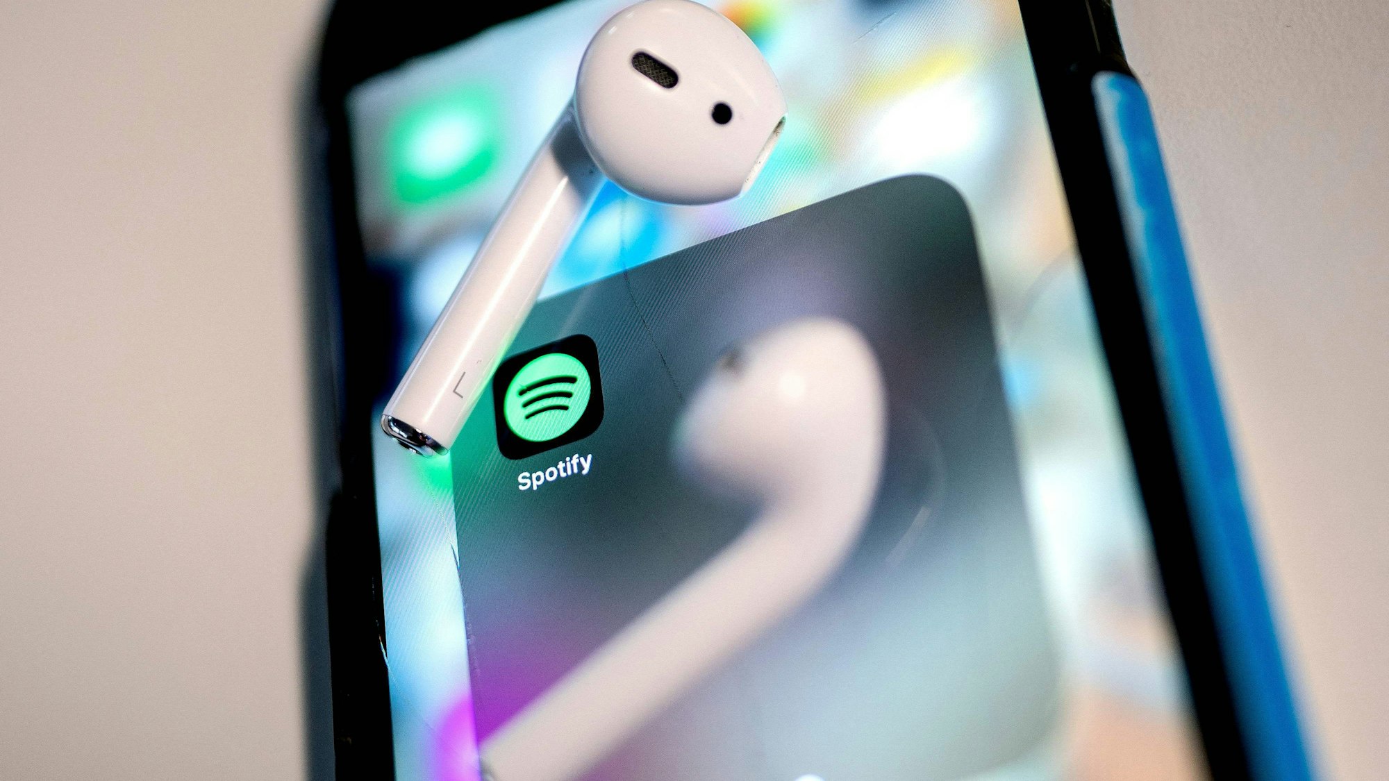 Auf dem Bild ist das Icon der Spotify-App auf einem Smartphone zu sehen. Auf dem Bildschirm liegen zwei kabellose Ohrhörer.