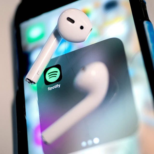 Auf dem Bild ist das Icon der Spotify-App auf einem Smartphone zu sehen. Auf dem Bildschirm liegen zwei kabellose Ohrhörer.&nbsp;
