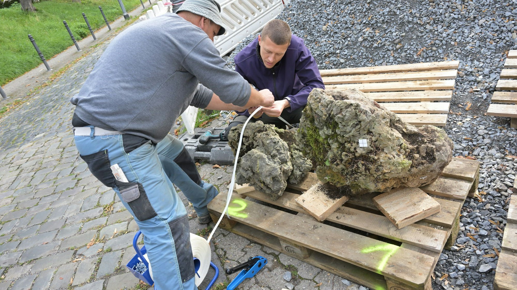 Zwei Arbeiter befestigen mit Kabelbinder einen kleinen und einen größeren Gesteinsbrocken auf einer Holzpalette.
