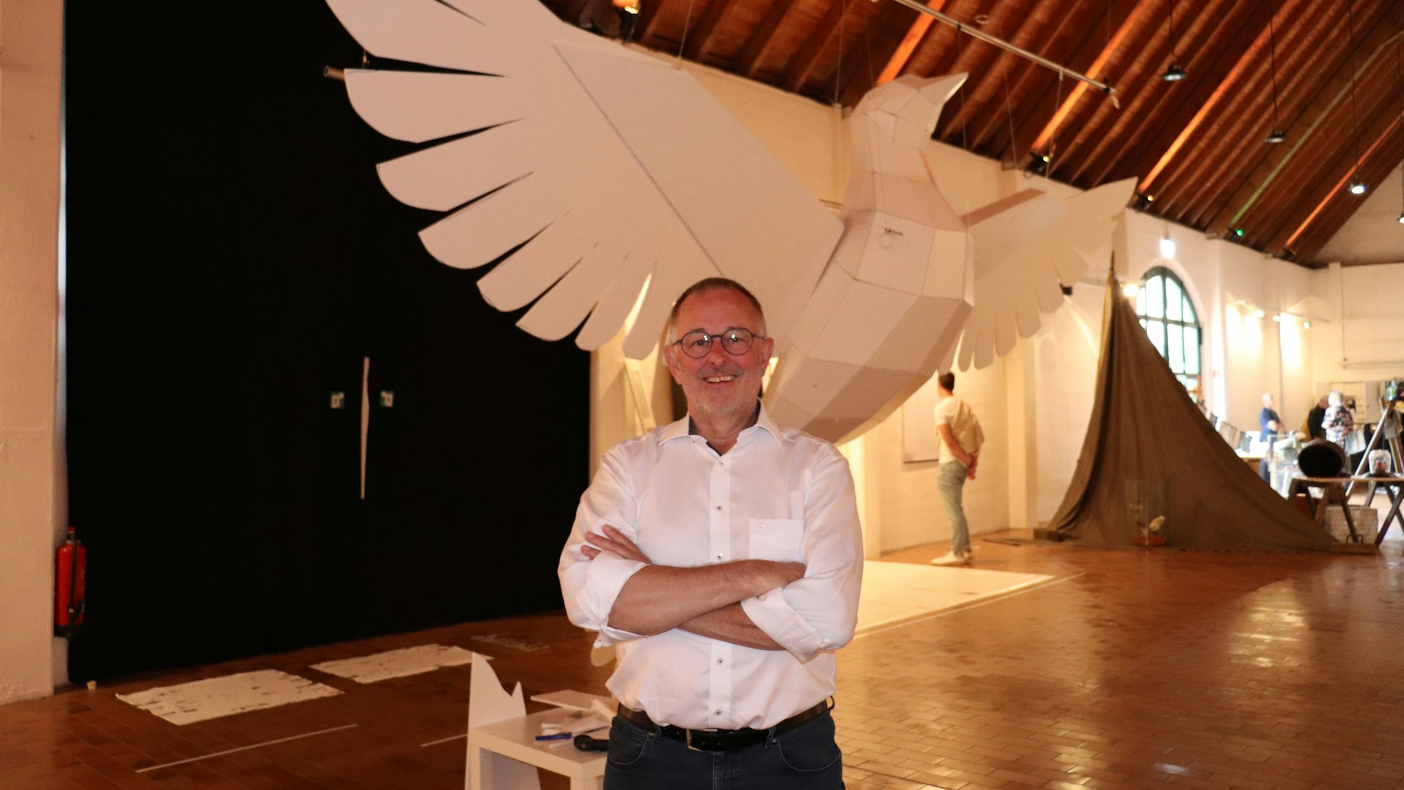 Eine riesige Taube aus Kunststoff hängt in einem Ausstellungsraum, davor steht der Künstler Manni Vogel, der sie gebaut hat.