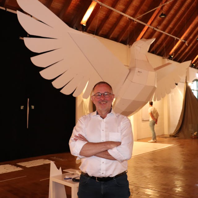 Eine riesige Taube aus Kunststoff hängt in einem Ausstellungsraum, davor steht der Künstler Manni Vogel, der sie gebaut hat.