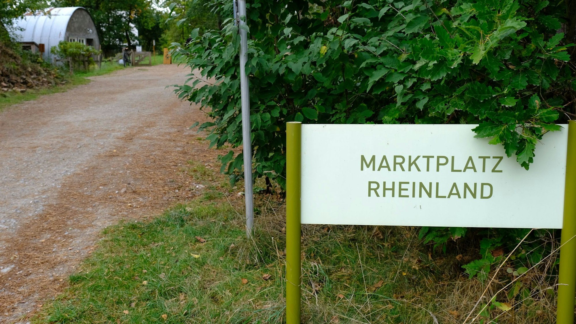 Ein Schild mit der Aufschrift Marktplatz Rheinland im LVR-Freilichtmuseum Kommern. Im Hintergrund sind sogenannte Nissenhütten zu sehen.