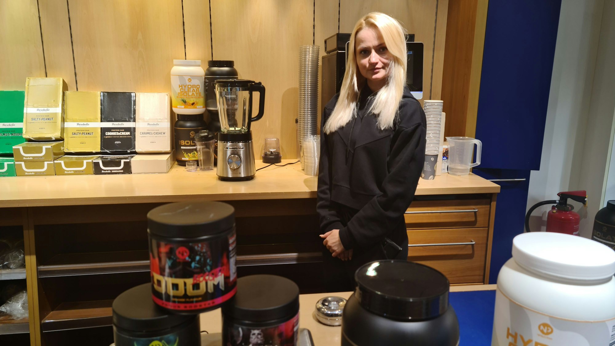 Eine Frau mit blondem langen Haar steht hinter einem Verkaufstresen, auf dem Behälter mit Protein-Pulver stehen.