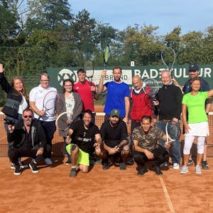 Eine Gruppe Menschen posiert in Sportkleidung und mit Tennisschlägern vor einem Tennisnetz.