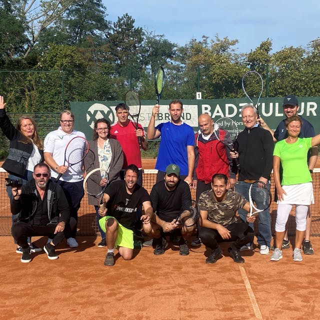 Eine Gruppe Menschen posiert in Sportkleidung und mit Tennisschlägern vor einem Tennisnetz.