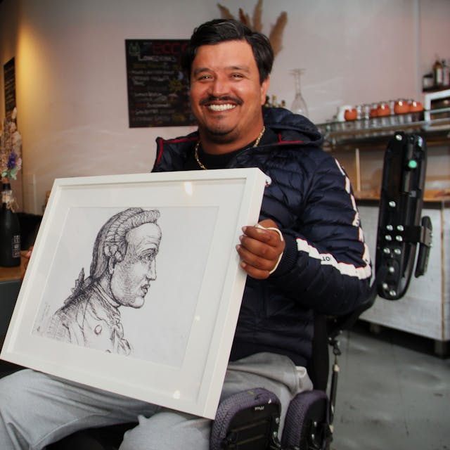 Ein dunkelhaariger sitzt in einem Rollstuhl, auf dem Schoß hält er ein Schwarz-Weiß-Porträt.