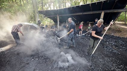 Frauen, Männer und Kinder arbeiten an einem Kohlenmeiler, um sie herum ist Rauch.