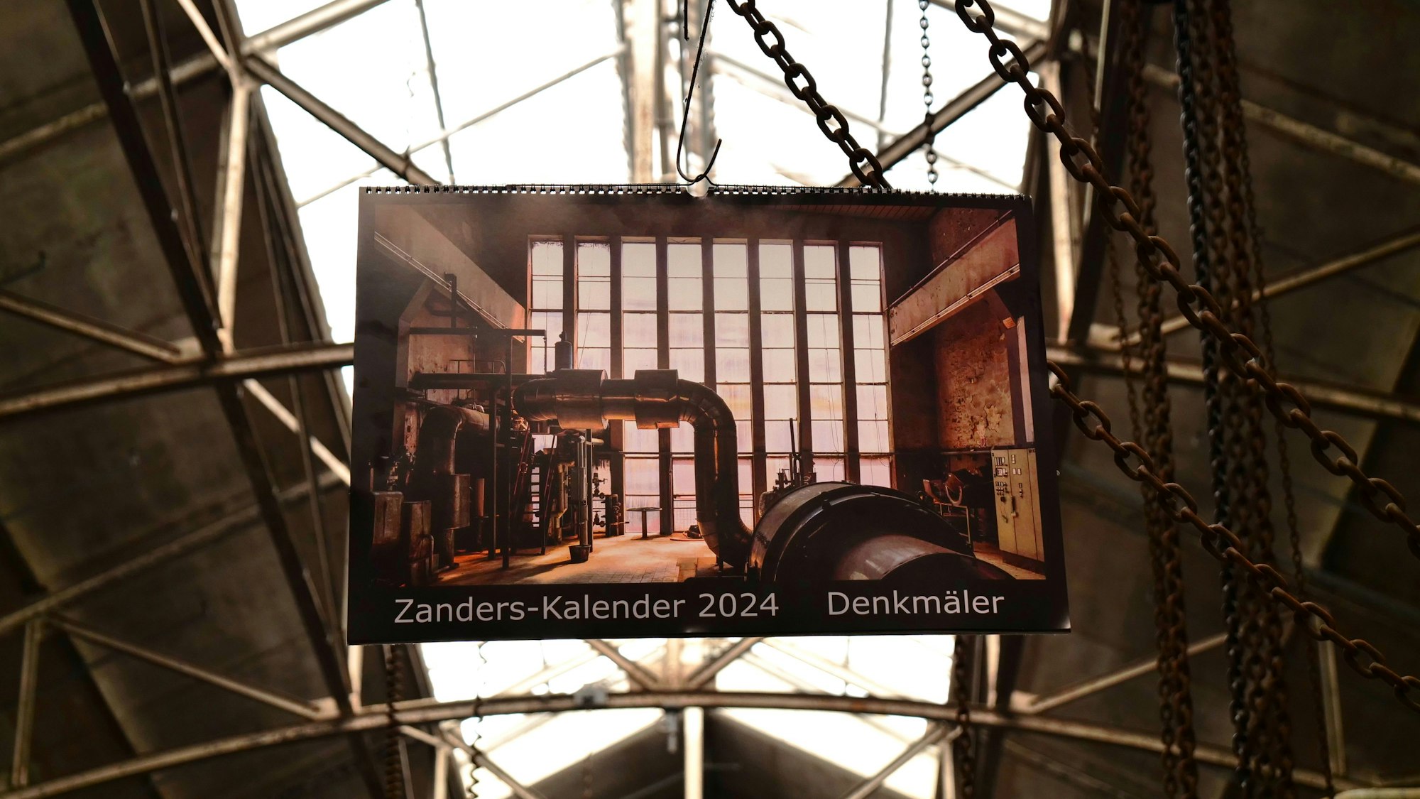 Ein Kalender mit der Aufschrift „Zanders-Kalender 2024“ hängt an einer Kette in einer alten Industriehalle.