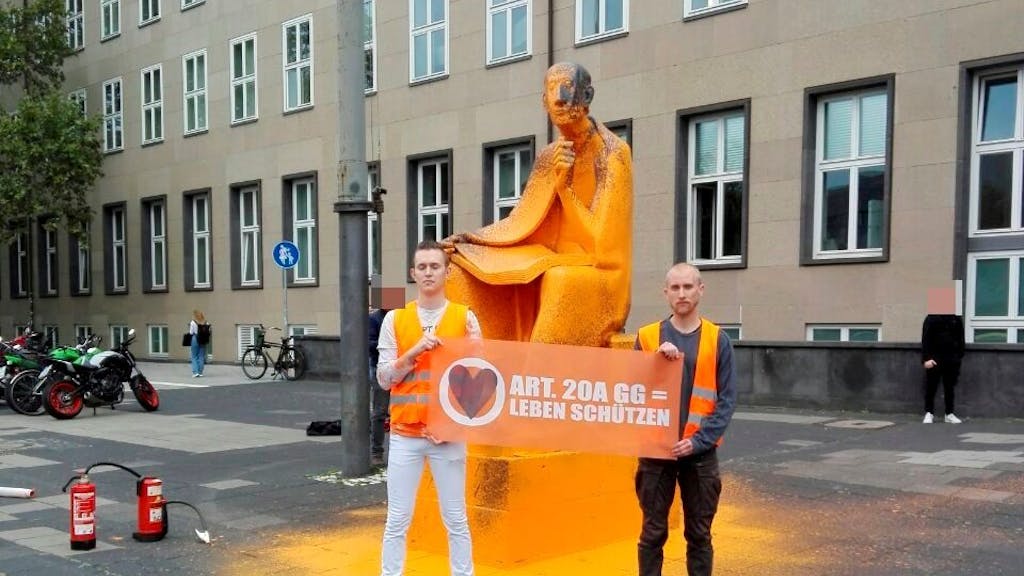 Zwei junge Männer stehen mit einem Plakat vor einem Denkmal, über das Farbe gegossen wurde.&nbsp;