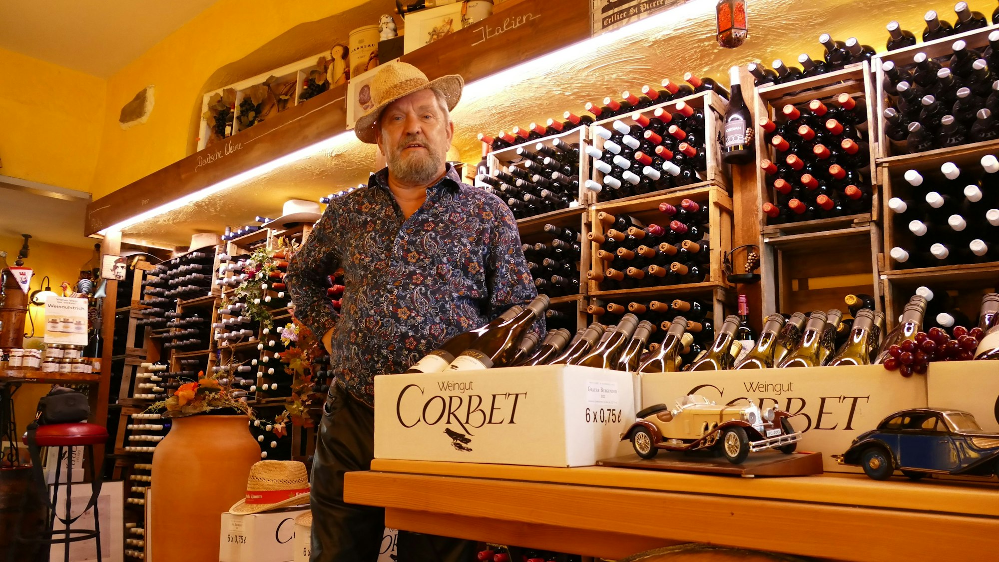 Ein Mann mit Hut und buntem Hemd steht vor einem Regal mit Weinflaschen.