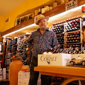Ein Mann mit Hut und buntem Hemd steht vor einem Regal mit Weinflaschen.