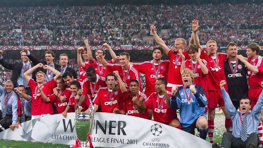 Die Bayern-Spieler stehen hinter dem Champions League Pokal und jubeln. im Hintergrund sind die Tribünen des San Siro in Mailand zu sehen.