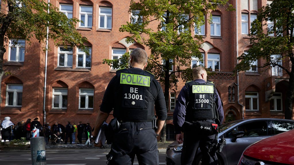 Zwei Polizisten stehen vor einer Berliner Schule. Im Hintergrund sind einige Jugendliche zu sehen.
