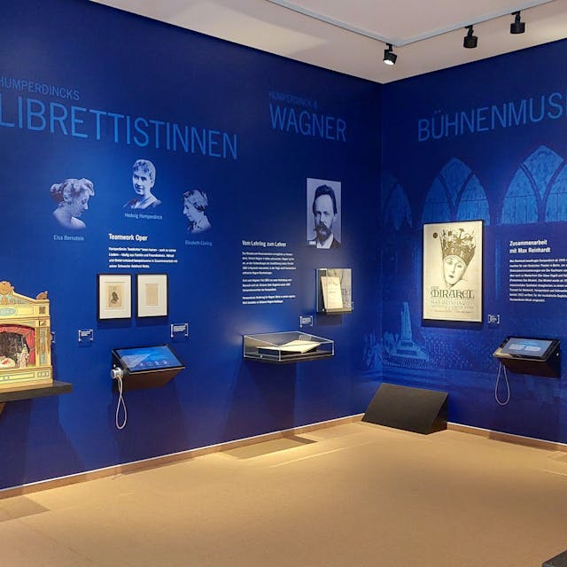 Neue Humperdinck-Abteilung im Stadtmuseum Siegburg. Zu sehen ist die Ausstellung. An großen blauen Wänden stehen Texte und Bilder, etwa der Librettistinnen seiner Stücke. An den Wänden hängen auch einige Bildschirme mit Kopfhörern.&nbsp;