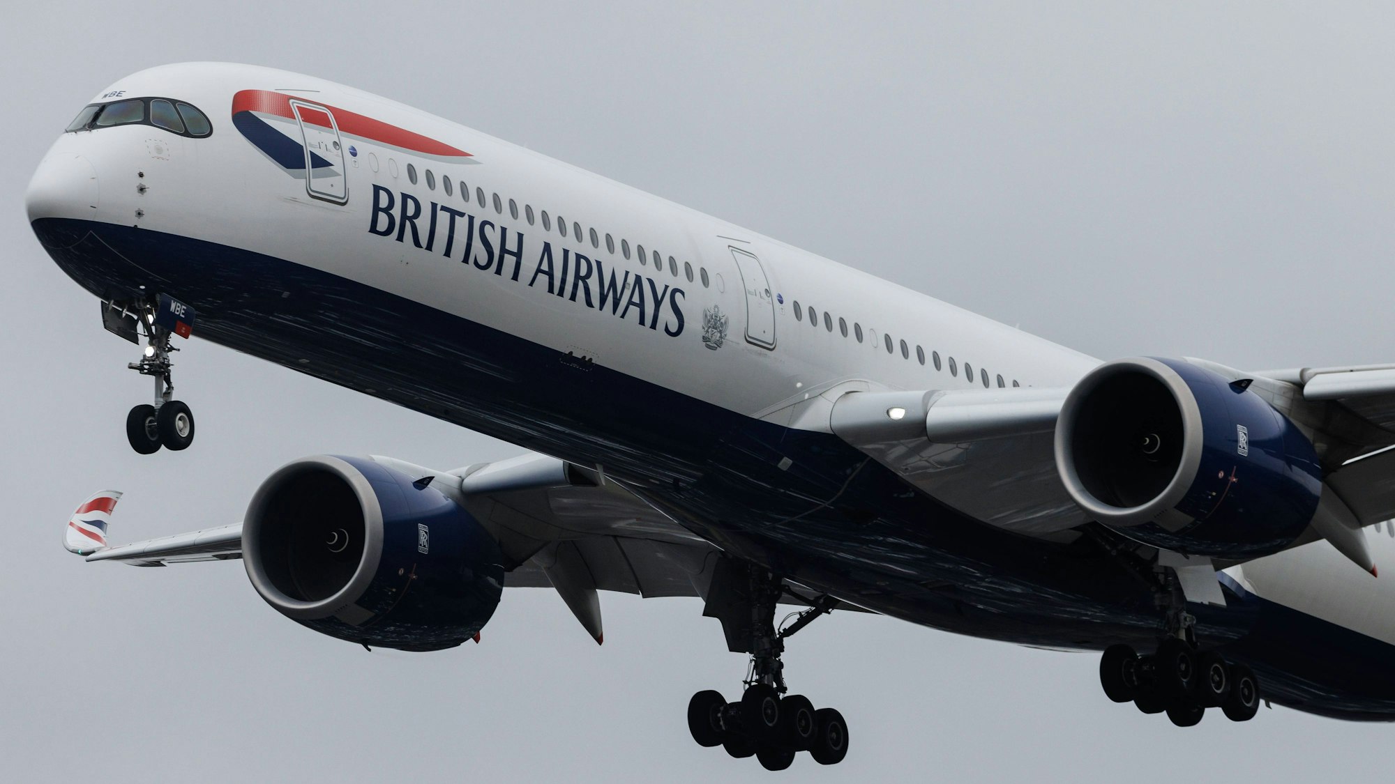 Ein Airbus A350 der britischen Fluggesellschaft British Airways ist mit ausgefahrenem Fahrwerk am Himmel zu sehen. Die Maschine ist blau-weiß-rot markiert.