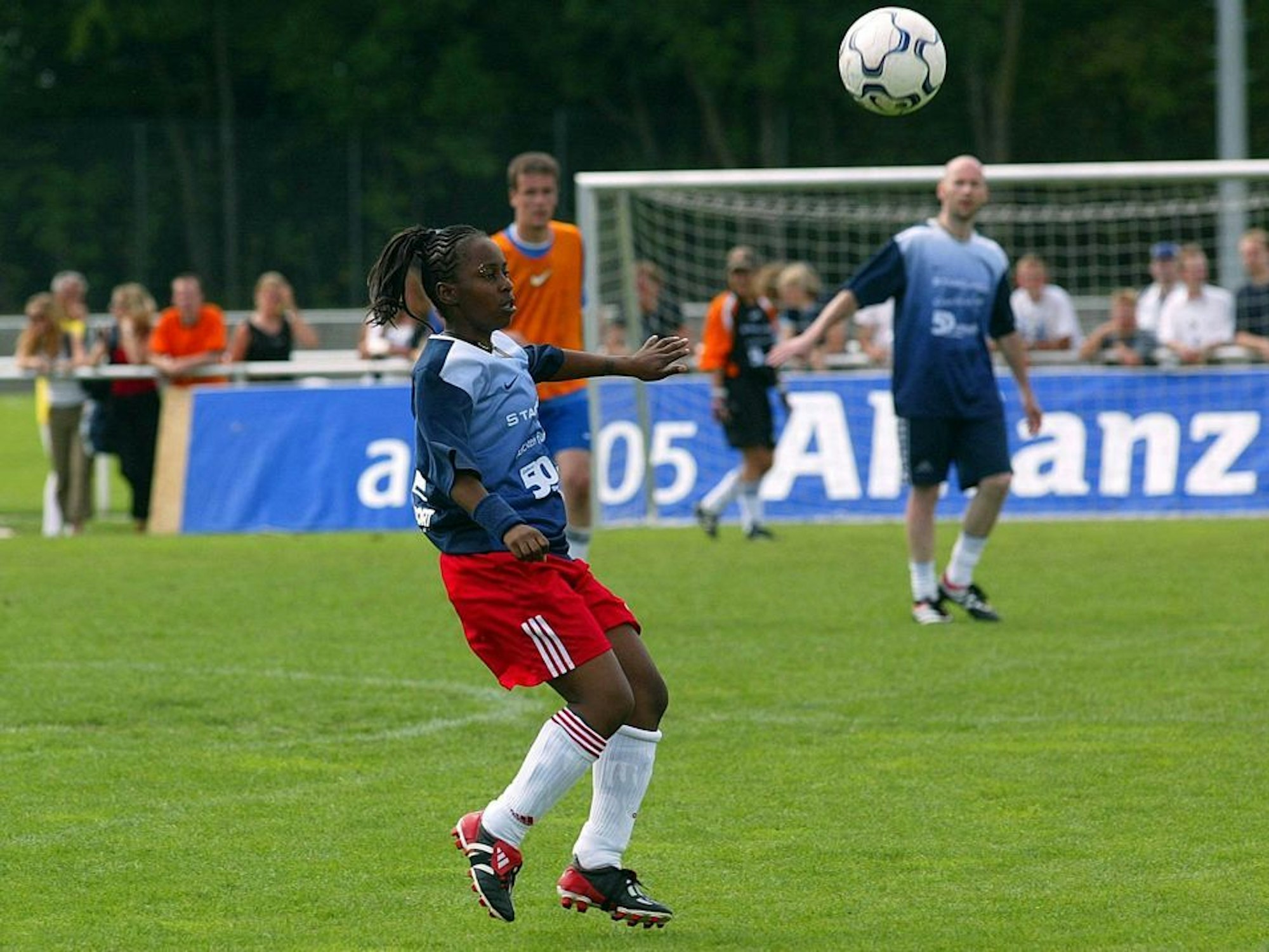 Eine Fußballerin spielt einen Ball in der Luft, im Hintergrund sind zwei Männer zu sehen, die gleiche Trikots tragen.