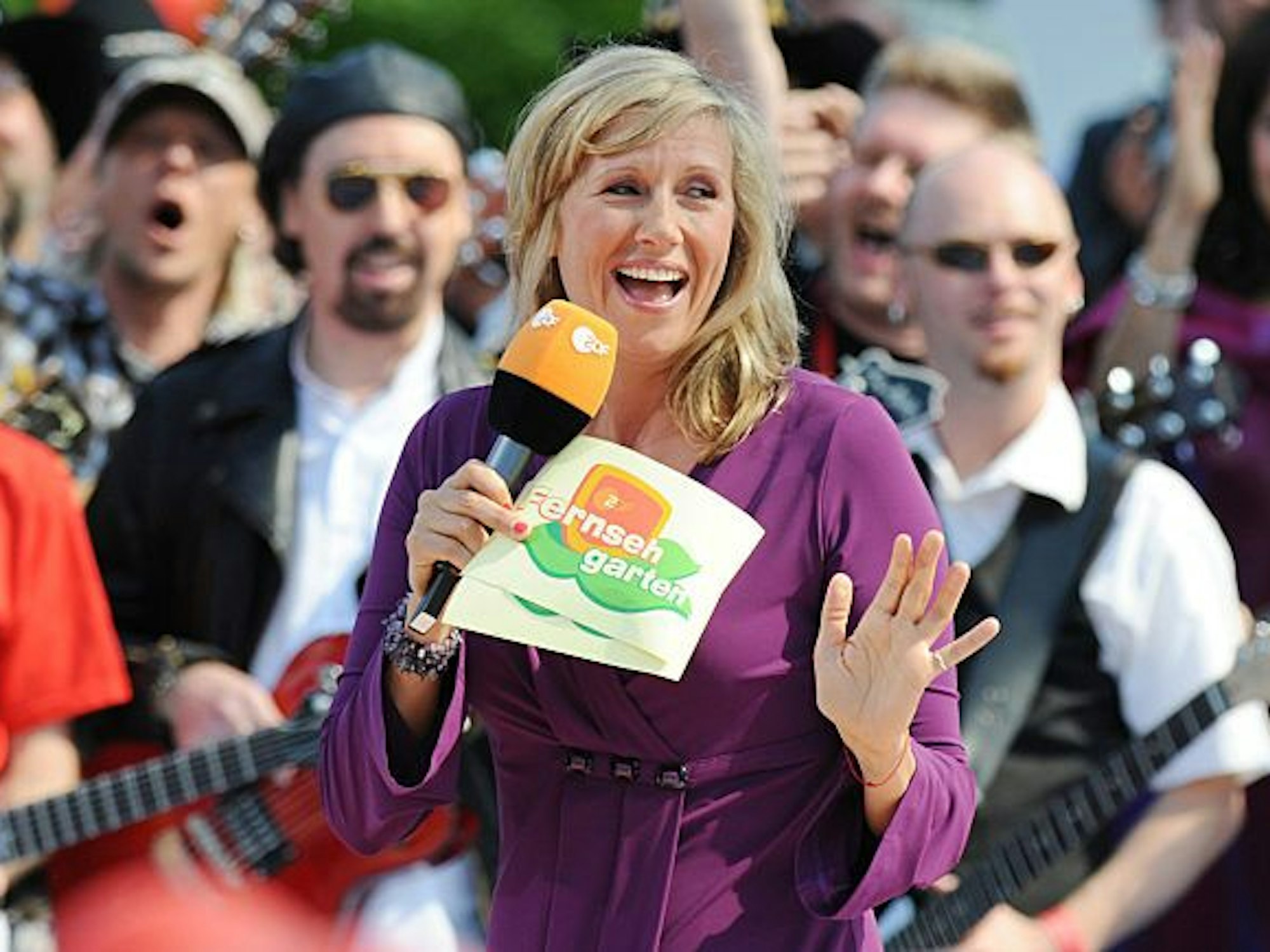 Andrea Kiewel im violetten Kleid. Sie lacht, hält ein ZDF-Mikrofon und Notizkarten vom ZDF-Fernsehgarten. Im Hintergrund steht eine Band.