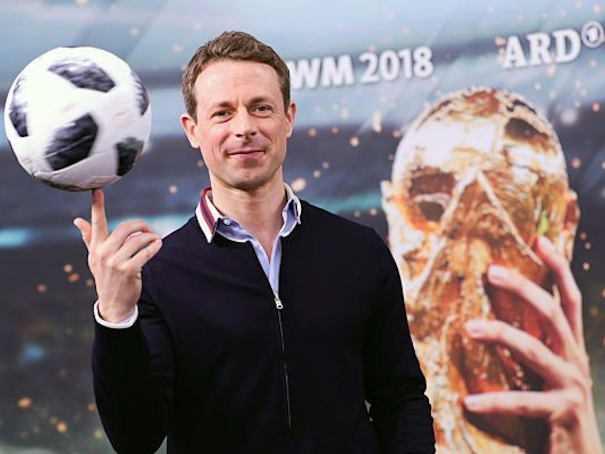 Alexander Bommes balanciert einen Fußball auf seinem Finger und steht vor einer Werbetafel der ARD zur Fußball-WM 2018.