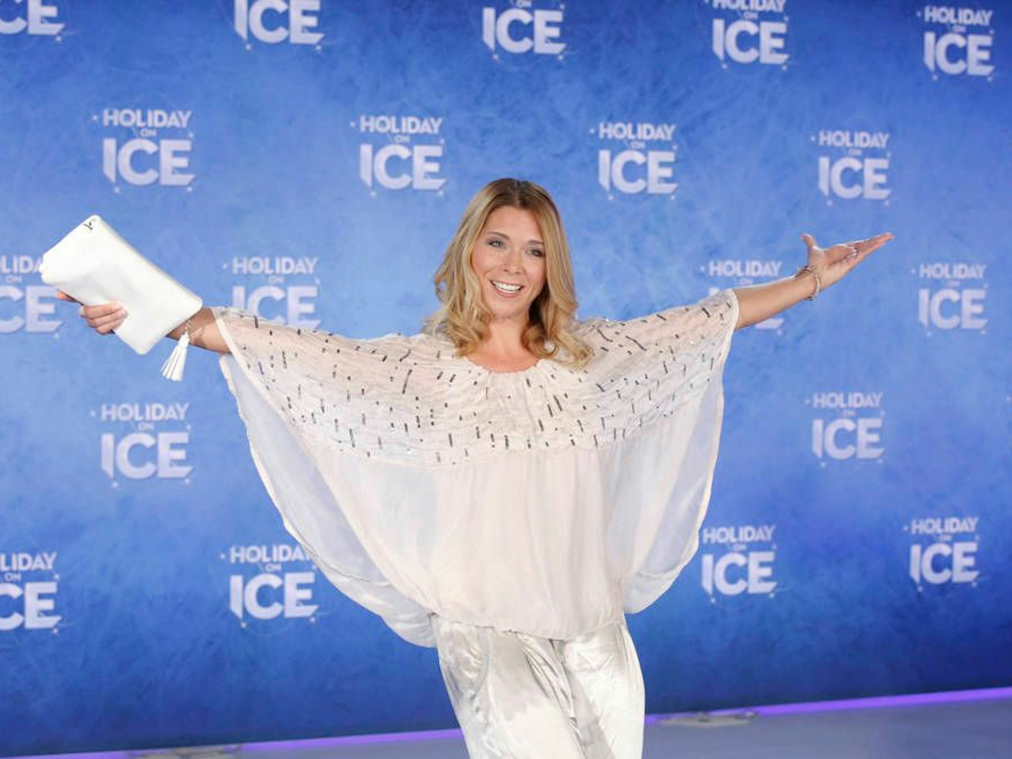 Tanja Szewczenko posiert vor einer Werbewand von „Holidays on Ice“ im weißen Outfit.