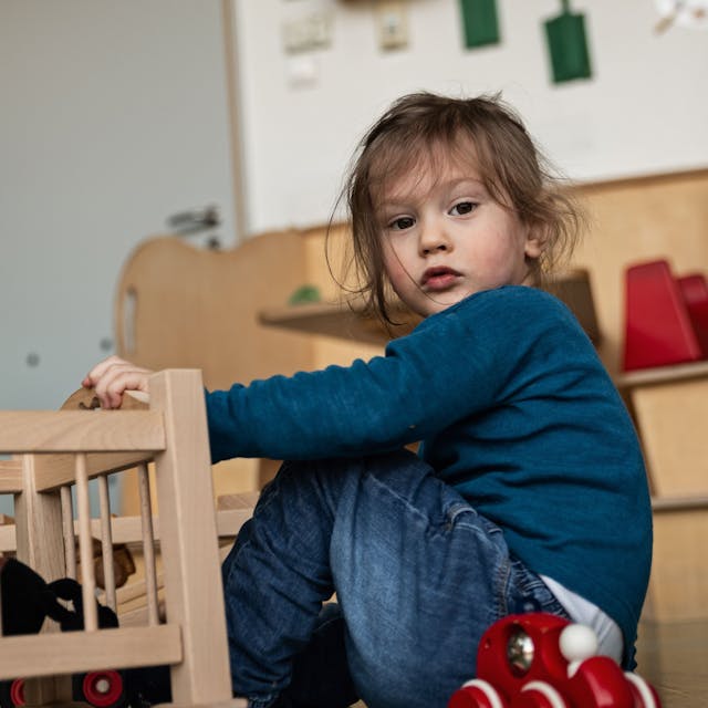 Ein Kind sitzt auf dem Boden neben einem Puppenbett und schaut in die Kamera.