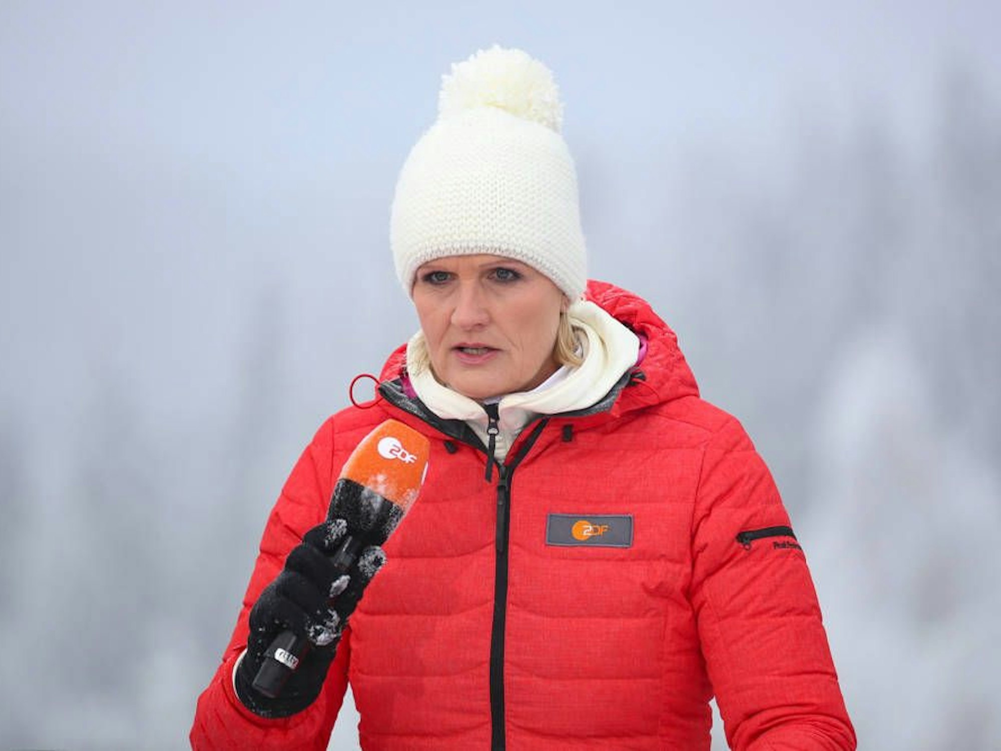 Kristin Otto steht in roter Winterjacke und weißer Pudelmütze mit einem ZDF-Mikrofon in einer Winterlandschaft.