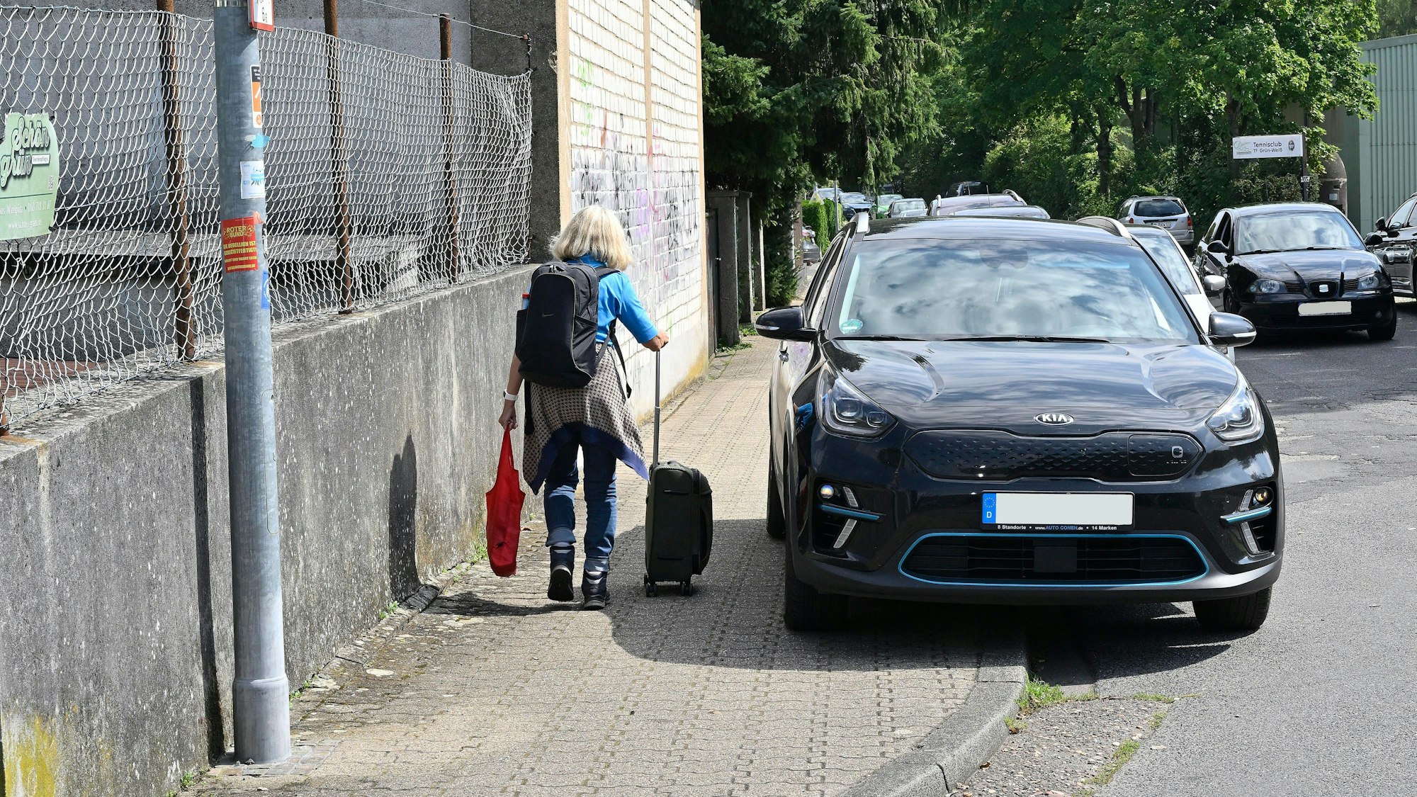 Eine Frau geht mit einem Rollkoffer vorbei an einem Auto, das zur Hälfte auf dem Gehweg steht.