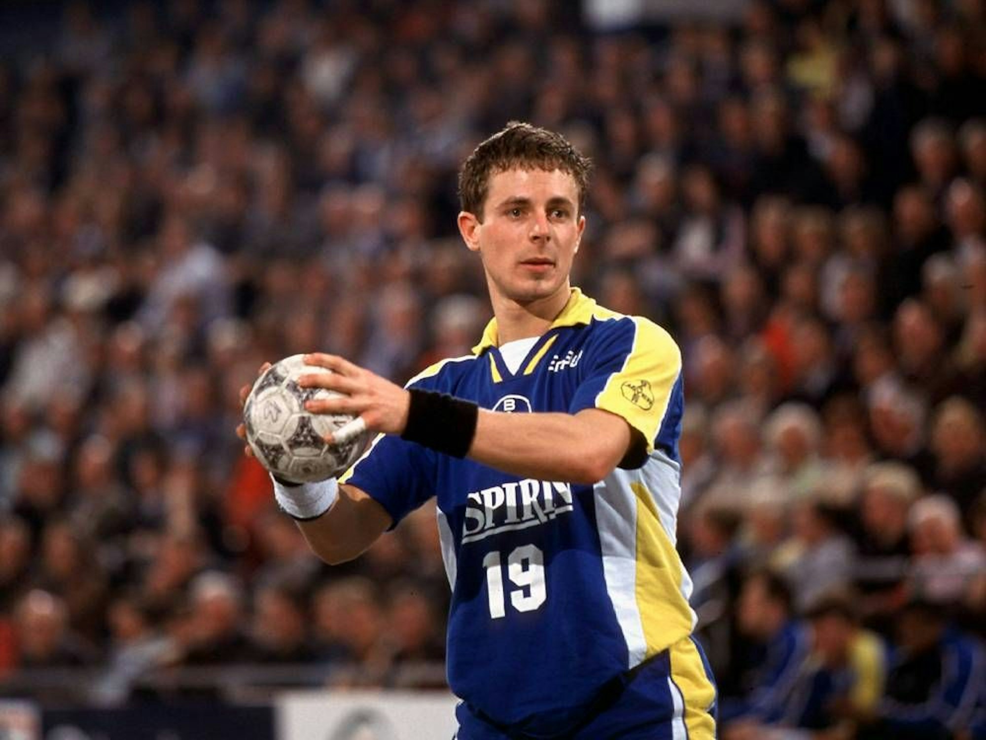 Ein junger Handballer hält den Ball im Stand bereit zum Wurf. Er trägt ein gelb-blaues Trikot mit Aspirin als Hauptsponsor.