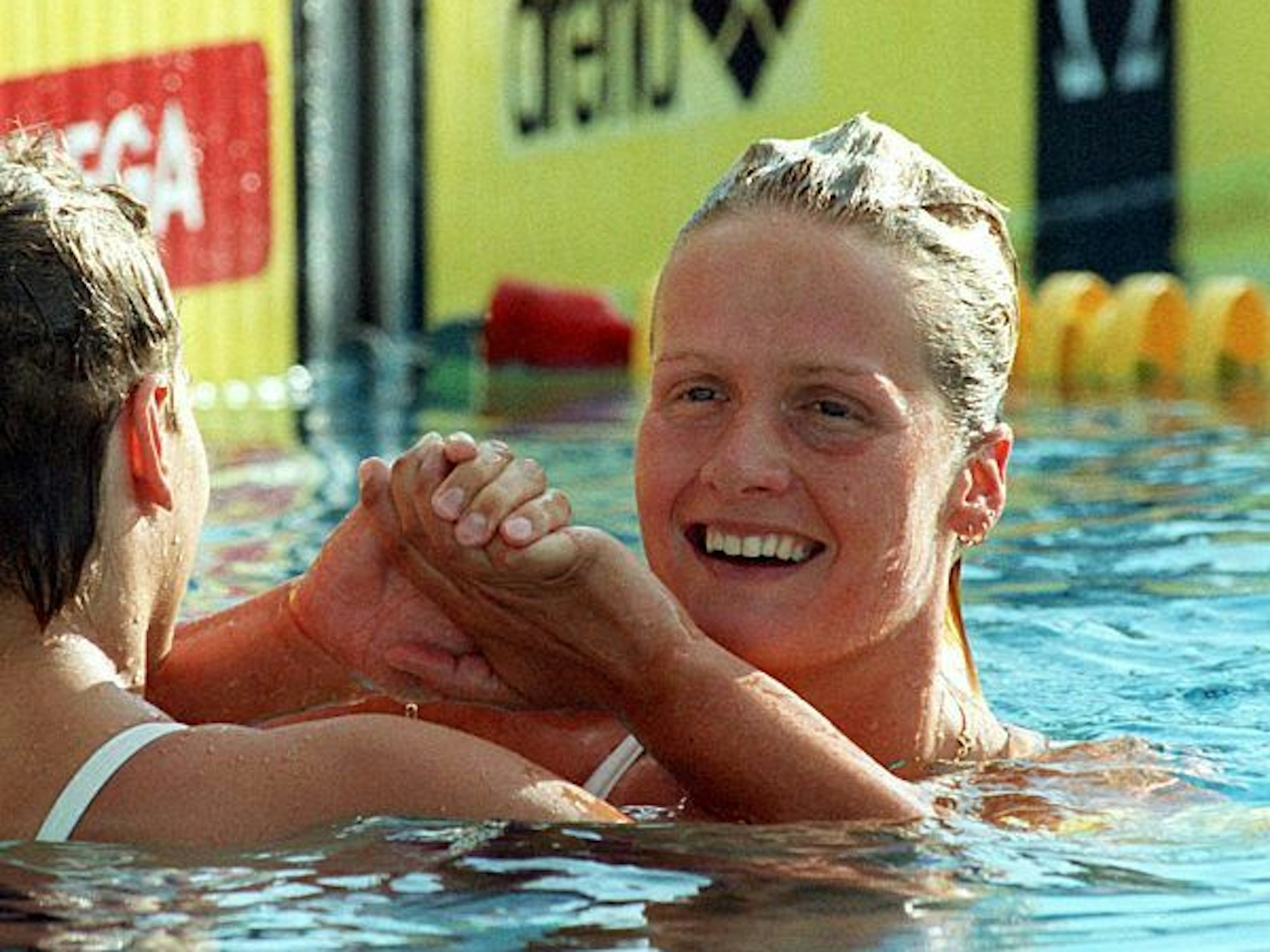 Eine junge Schwimmerin mit blonden Haaren im Becken. Sie lacht und klatscht mit einer anderen Schwimmerin ab.