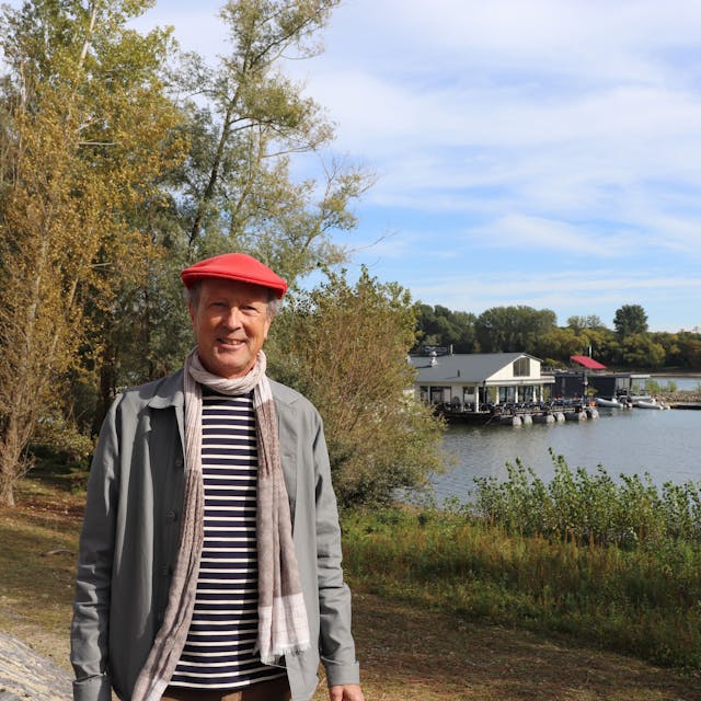 Ein Mann mit roter Baskenmütze auf dem Kopf steht am Rheinufer, im Hintergrund ist das Sürther Bootshaus zu sehen.