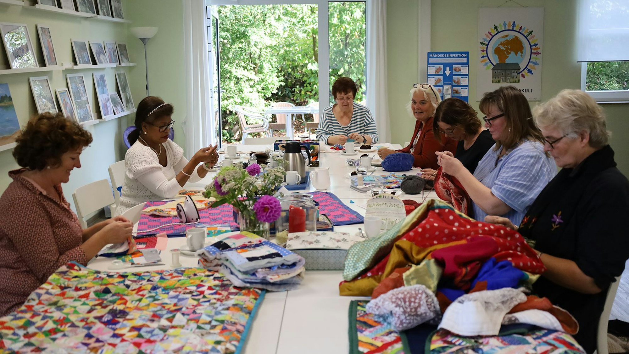 Sieben Frauen sitzen bei ihrer Handarbeit an einem großen Tisch, auf dem einige fertige Patchwork-Decken liegen.