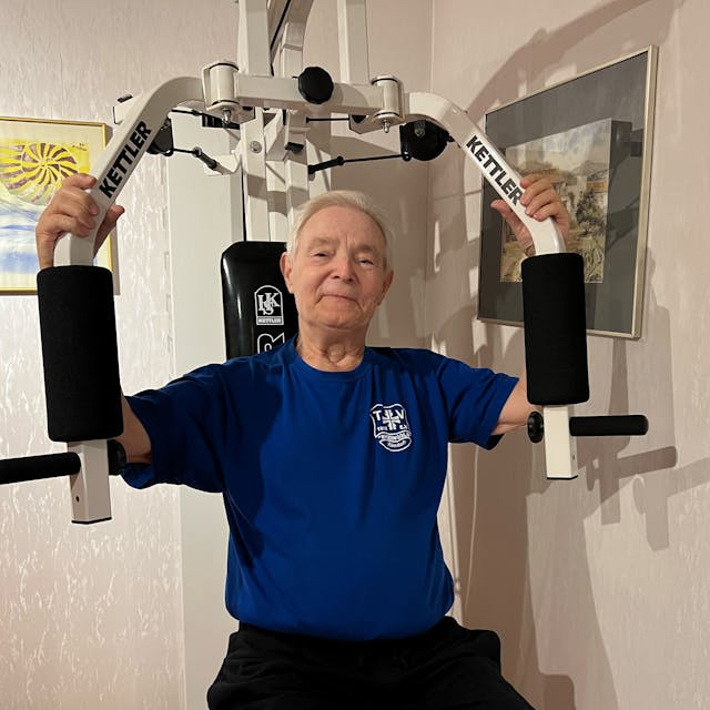 Ein älterer Mann mit grauem Haar sitzt auf einem Fitnessgerät, das die Armmuskulatur stärkt.