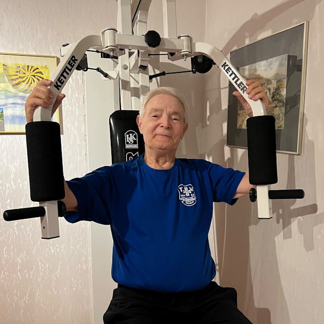 Ein älterer Mann mit grauem Haar sitzt auf einem Fitnessgerät, das die Armmuskulatur stärkt.
