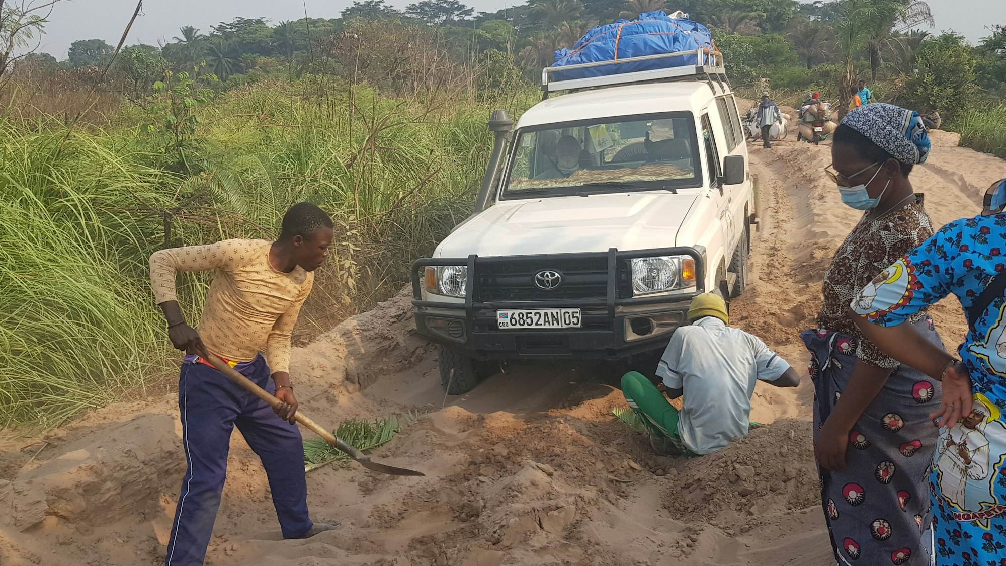 Der Wagen, mit dem Hans Jagsch und Manfred Janz durch den Kongo fuhren, steckt im Sand fest. Zwei Männer, einer mit Schaufel, graben das Fahrzeug frei.