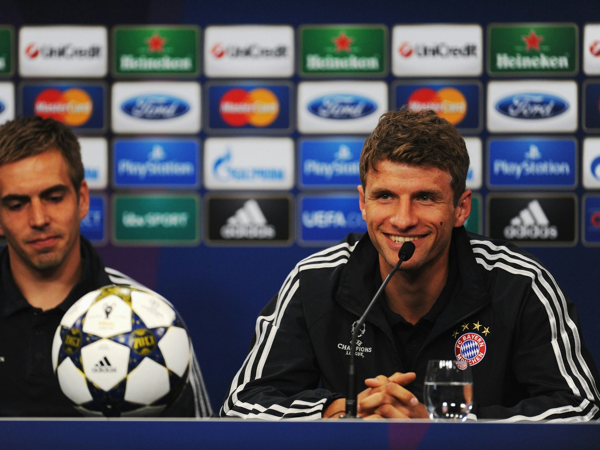 Thomas Müller grinst bei einer Pressekonferenz der Champions League. Im Hintergrund sitzt Kapitän Philipp Lahm.