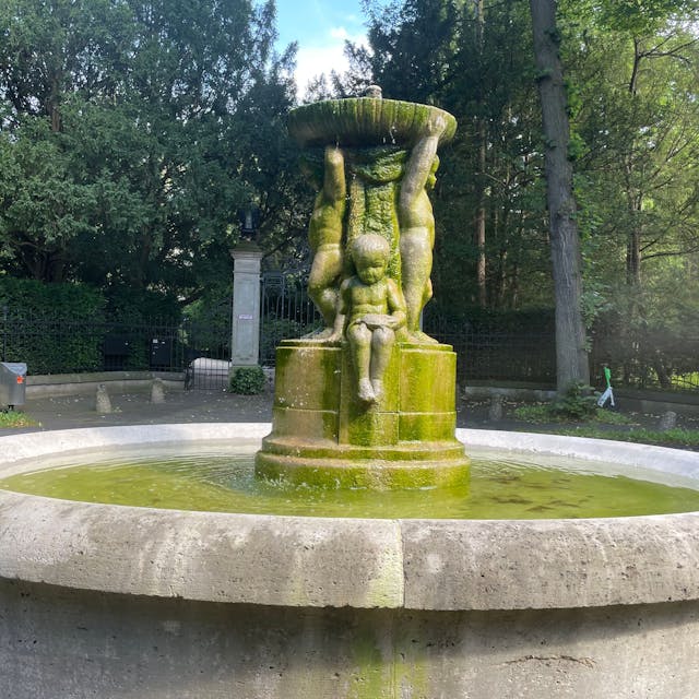 Ein steinerner Brunnen mit kindlichen Figuren steht in einer Parkanlage.