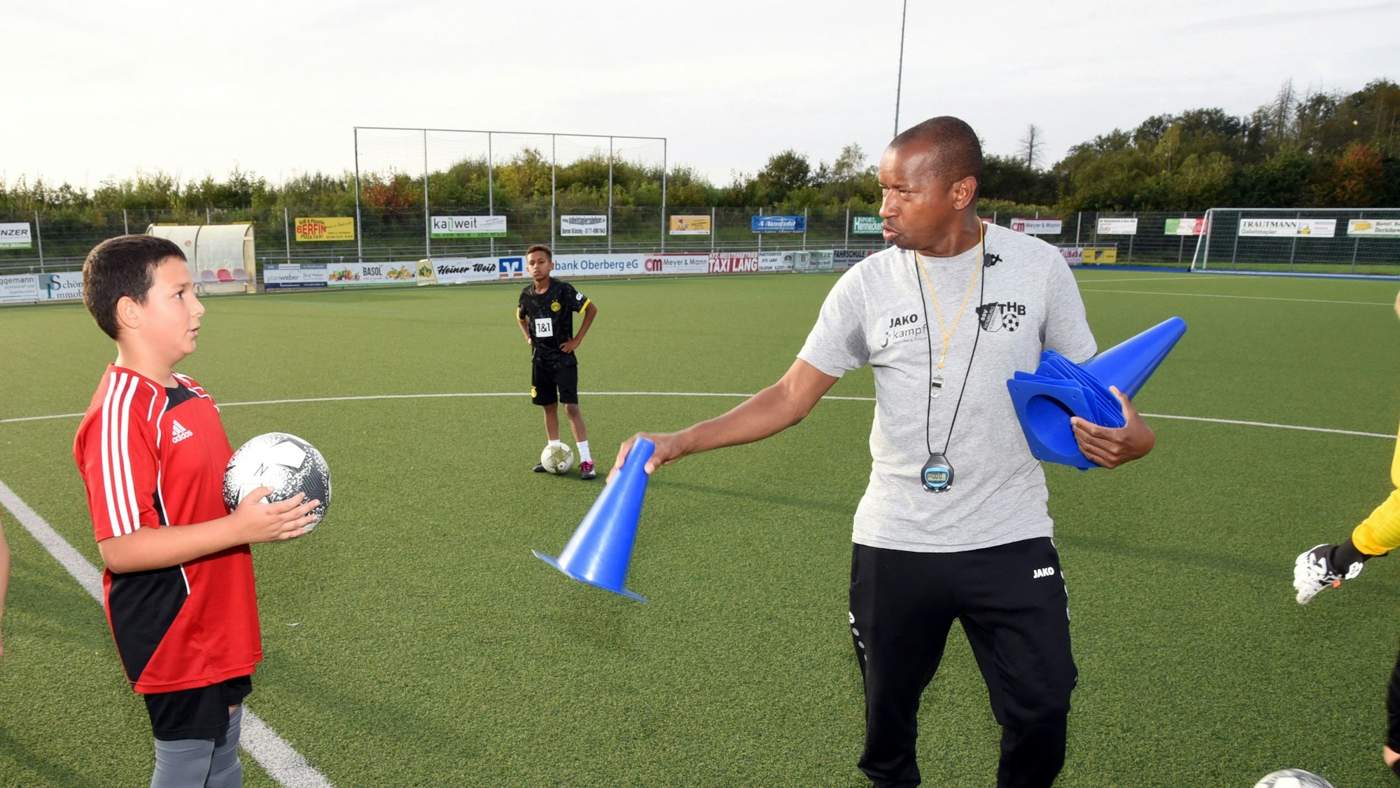 Abdoul Schwotzer Conde steht auf einem Fußballplatz und hält ein blaues Hütchen in der Hand, links von ihm steht ein Jugendlicher mit einem Fußball in den Händen.