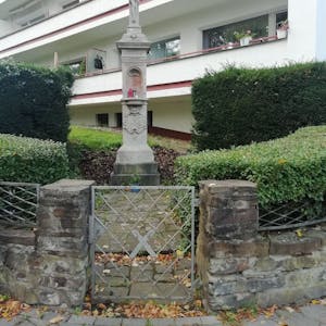 Ein steinernes Wegekreuz in einer Grünanlage vor einem Mehrfamilienhaus.