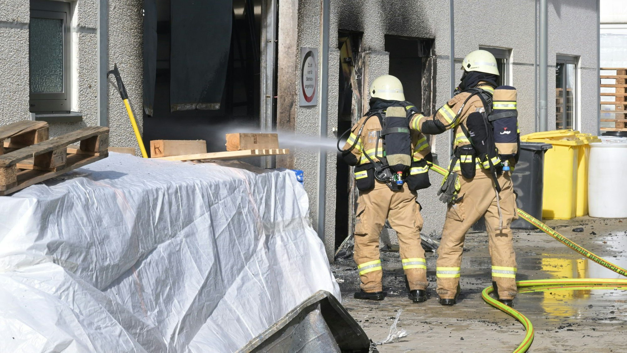 Zwei Feuerwehrleute halten einen Wasserschlauch in eine offenkundig brennende Lagerhalle.