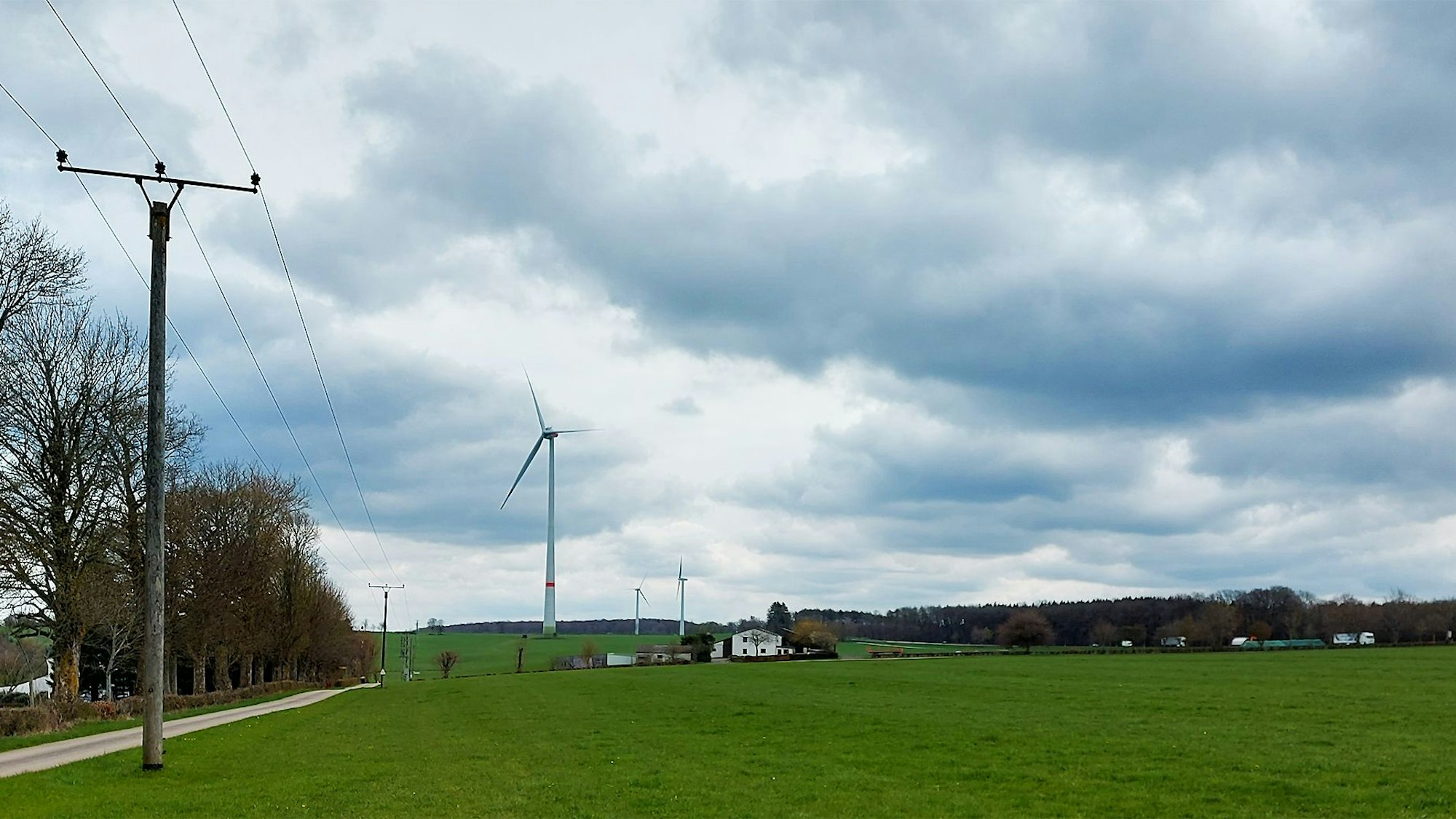 Auf einer Wiese sollen Photovoltaik-Module aufgebaut werden. Im Hintergrund sind ein Bauernhof und ein Windrad zu sehen, links eine Stromleitung.
