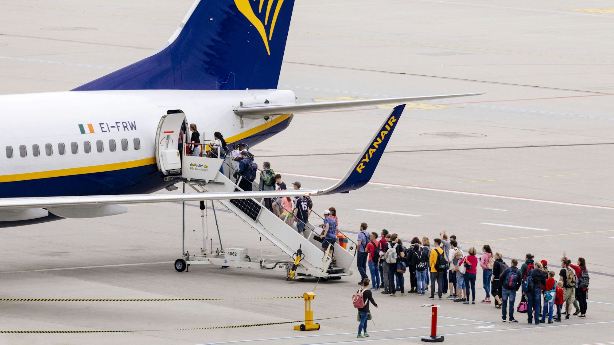 Diverse Menschen stehen am Flughafen Köln/Bonn vor einem Flugzeug und steigen über eine Außentreppe ein.