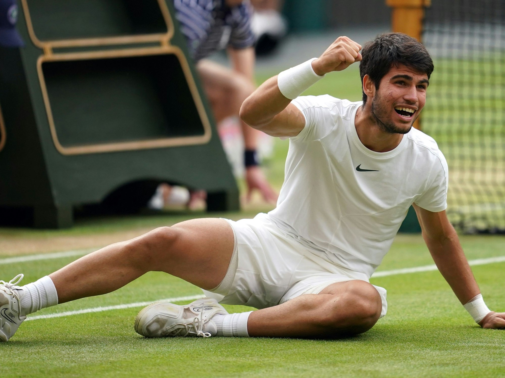 Carlos Alcaraz sitzt in Wimbledon auf dem Rasen und jubelt über seinen Titelgewinn.