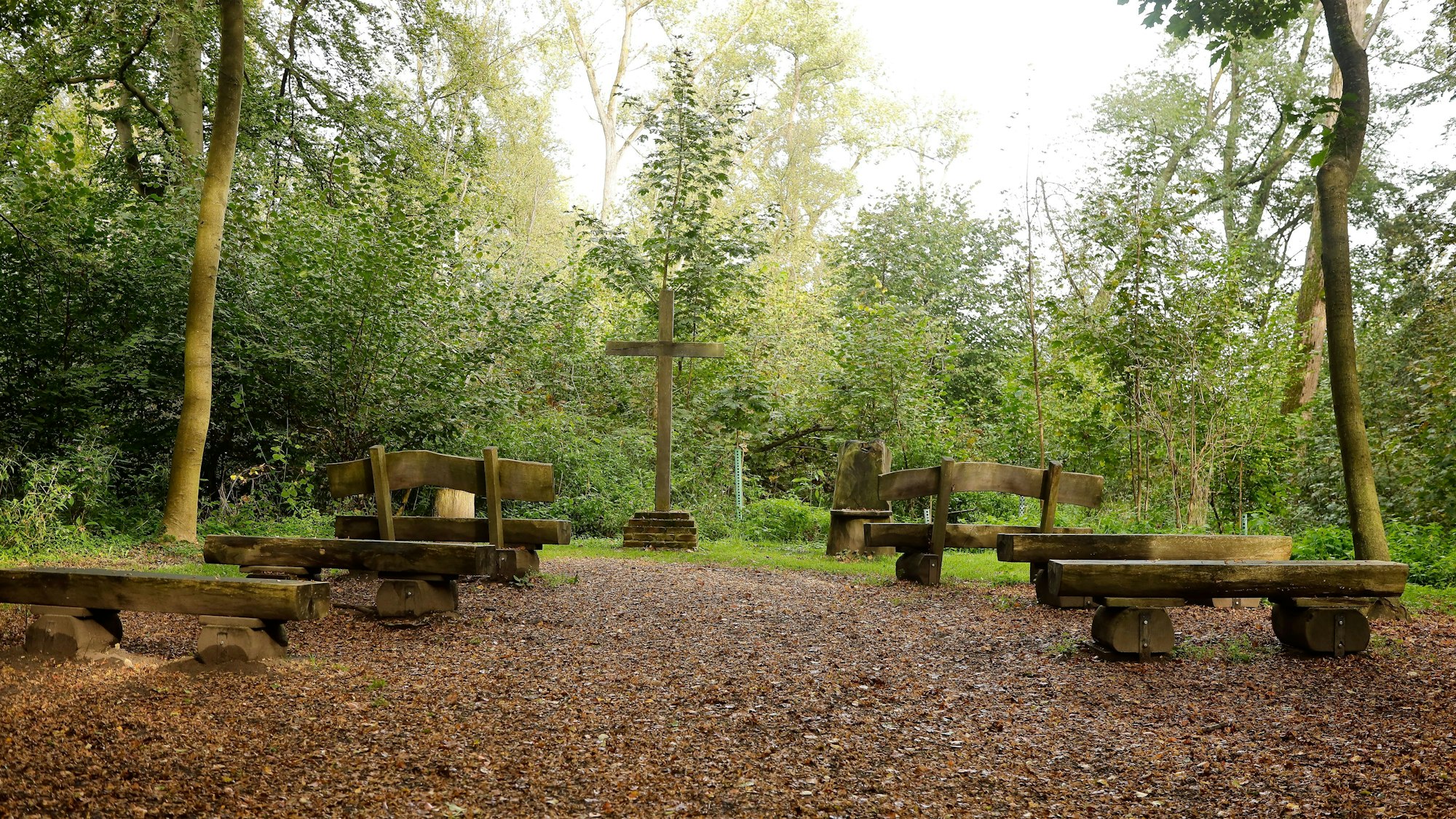 Mit Blick auf ein Holzkreuz sind einige individuelle Bänke aus Baumstämmen im Wald aufgestellt.