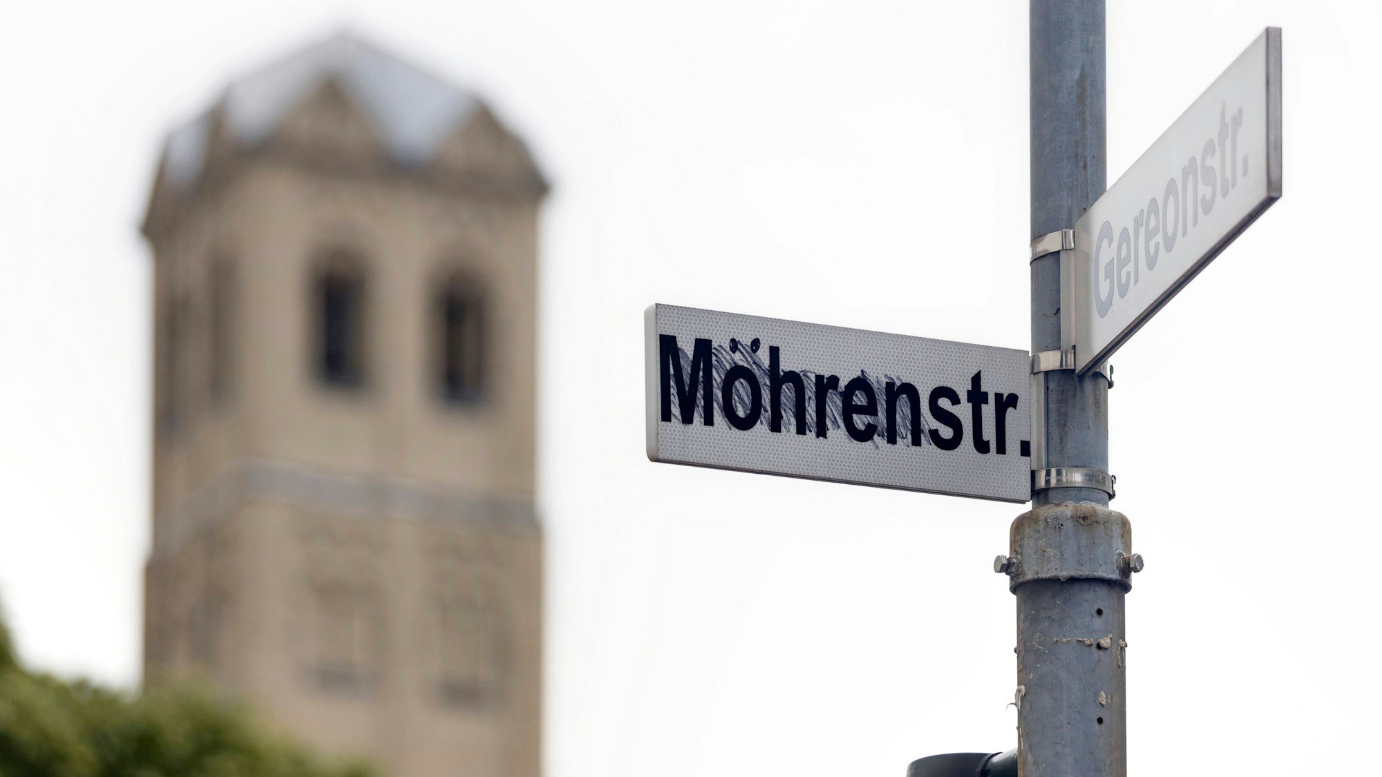 An einem Straßenmast sind zwei Straßenschilder, „Mohrenstraße“ und „Gereonstraße“, angebracht, wobei erstere von Unbekannten in „Möhrenstraße“ umgewandelt wurde. Im Bildhintergrund ist die Kirche Sankt Gereon zu sehen.