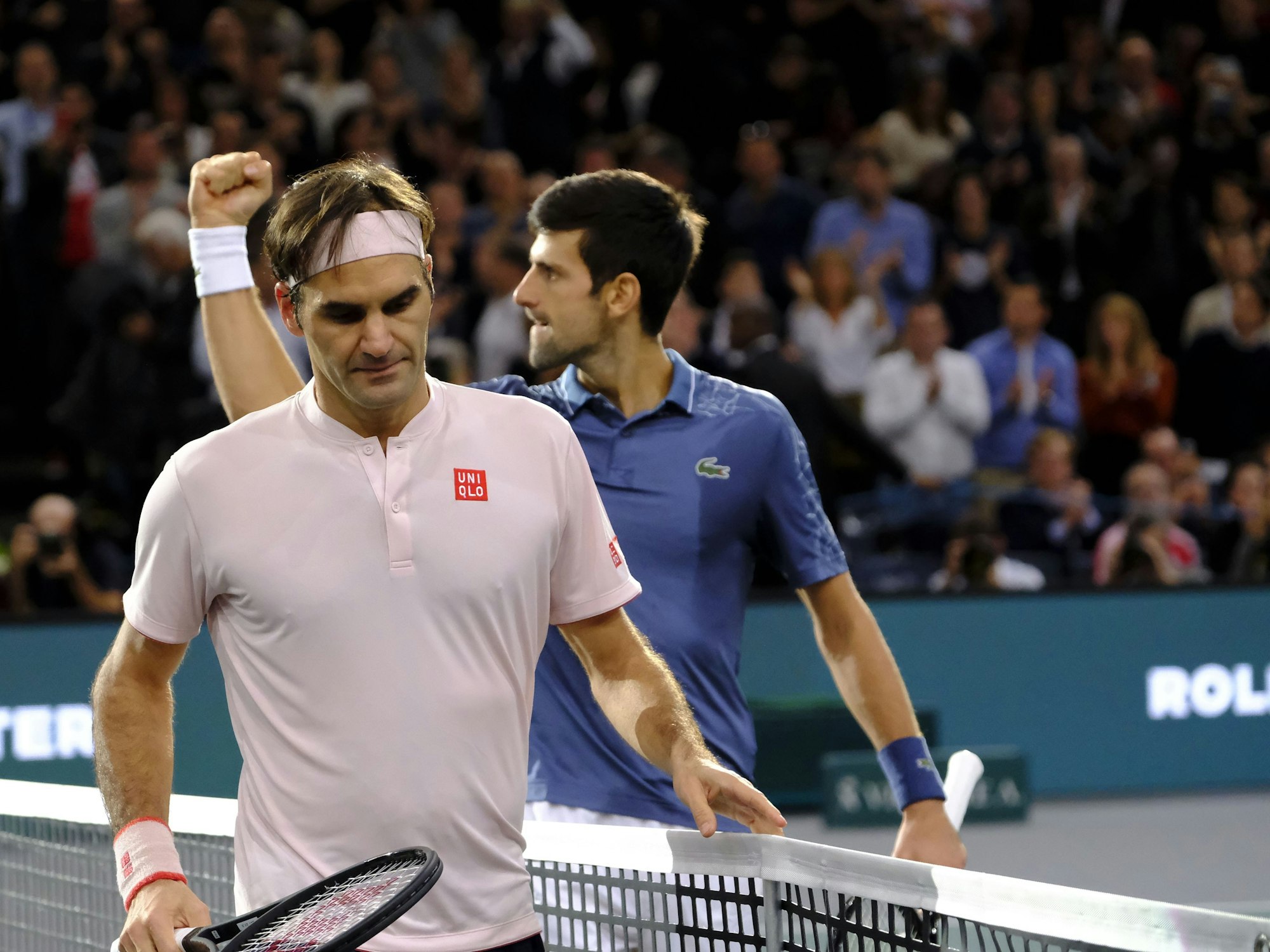 Roger Federer nach einem verlorenen Match enttäuscht am Netz, im Hintergrund jubelt Novak Djokovic.