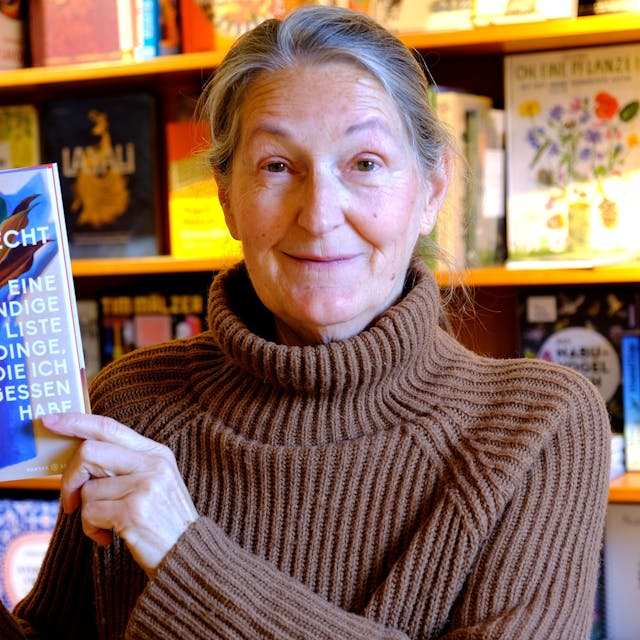 Iris Pütz steht vor einem Buchregal. Sie hat ein Exemplar eines Buchs von Doris Knecht in der Hand.