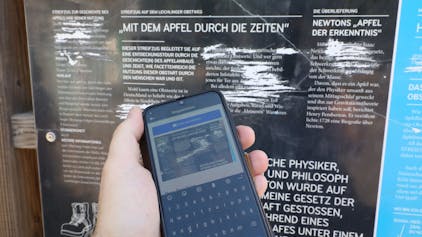 Vor einer zerkratzten Infotafel eines Bergischen Streifzug ist ein Handy zu sehen, mit dem gerade ein Foto für eine Meldung des Schadens aufgenommen wurde.