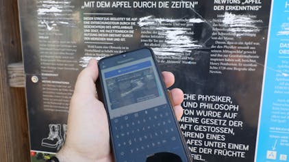 Vor einer zerkratzten Infotafel eines Bergischen Streifzug ist ein Handy zu sehen, mit dem gerade ein Foto für eine Meldung des Schadens aufgenommen wurde.