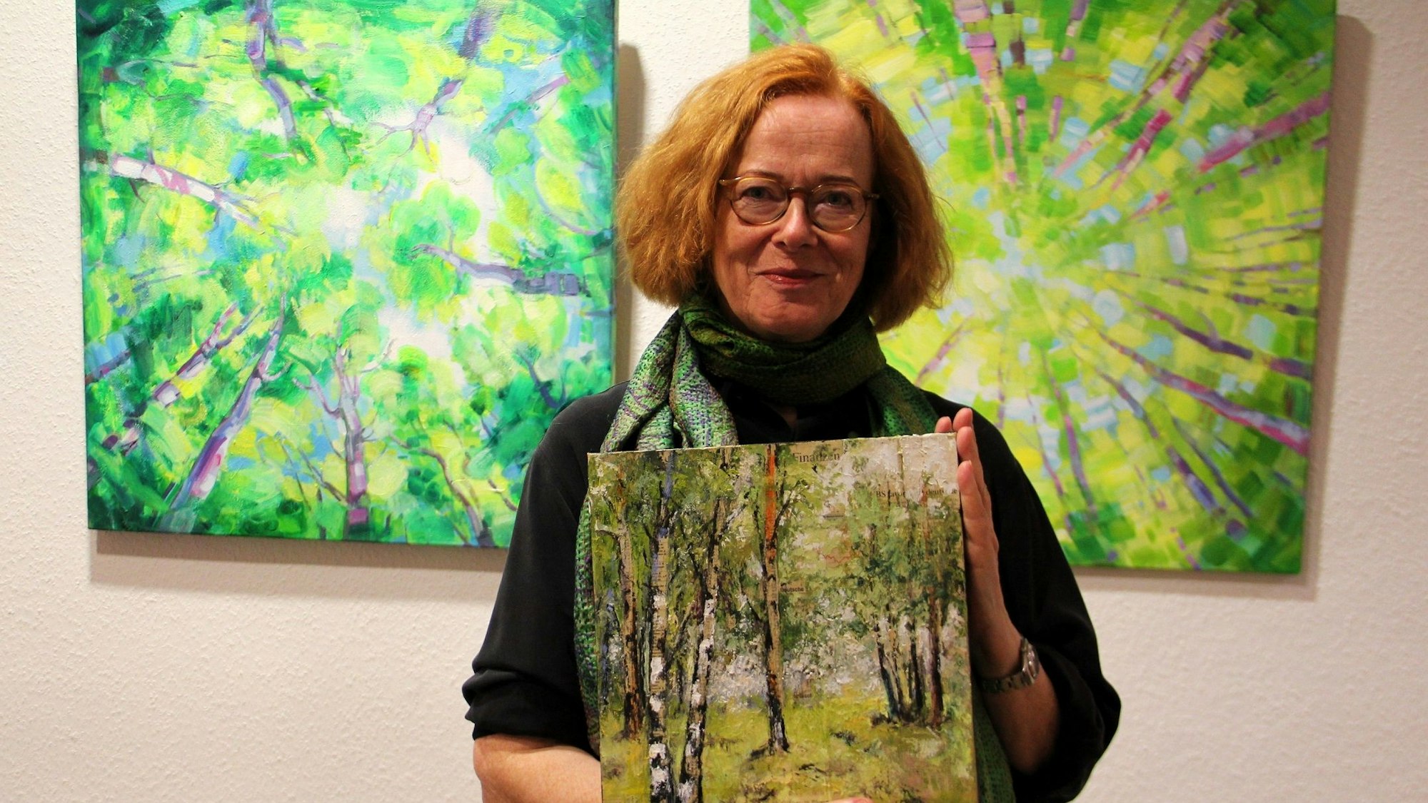 Eine Frau mit rotem Haar und Brille hält ein Bild von einem Wald in den Händen. Sie steht vor einer Wand, an der weitere Waldbilder von ihr hängen.