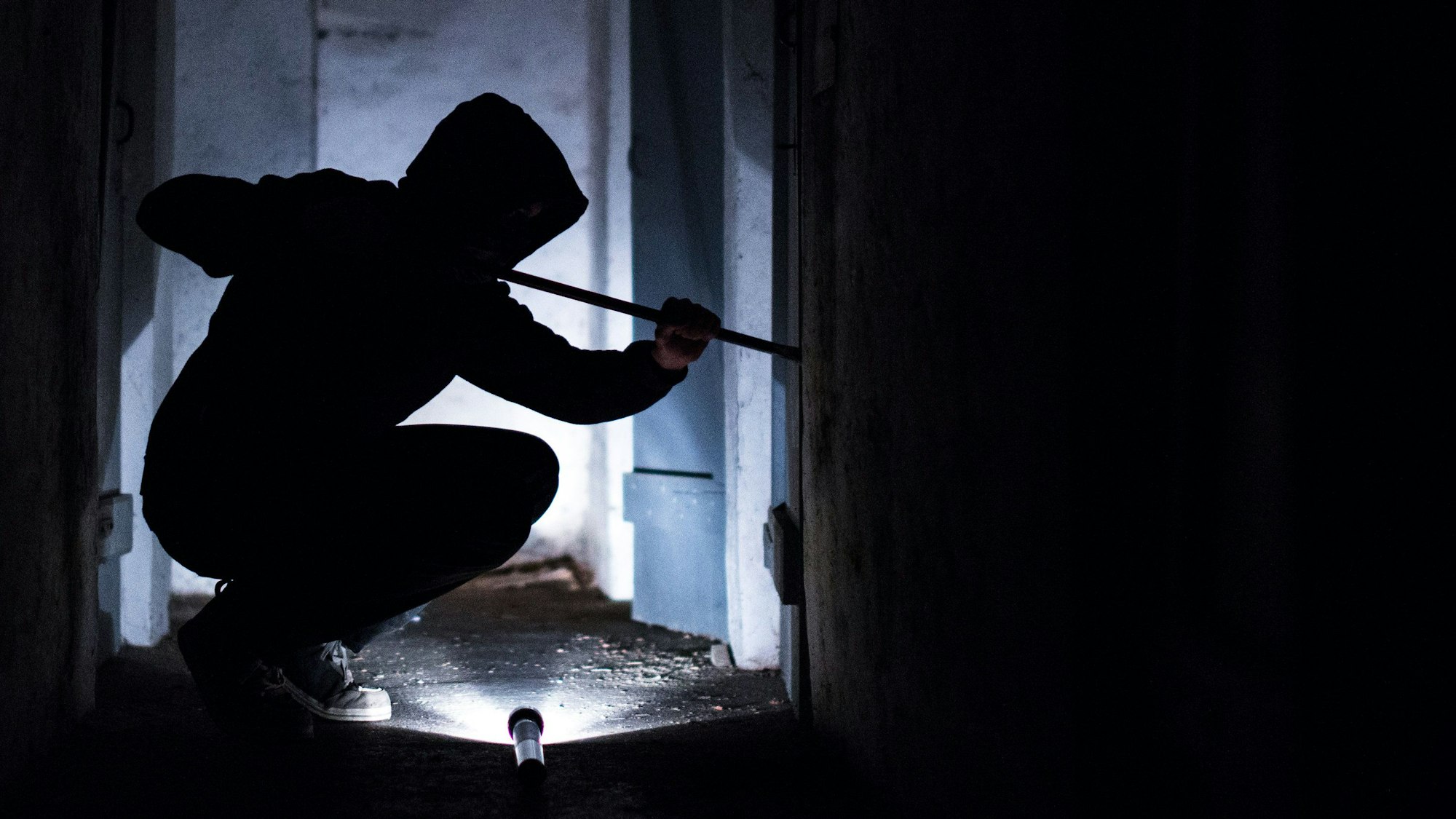 Ein Mann hebelt mit einem Brecheisen eine Tür im Keller eines Wohnhauses auf. (Symbolbild)