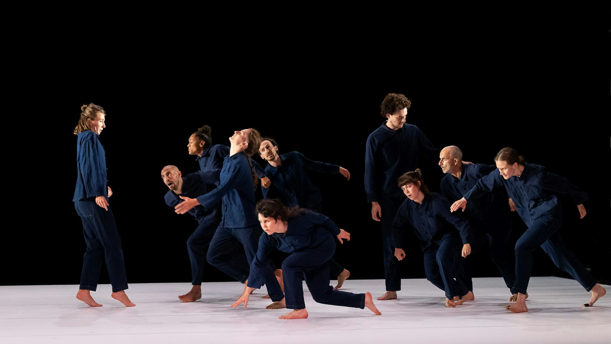 Die Tänzerinnen und Tänzer tragen blaue Hosen und Hemden und bewegen sich alle auf eine Tänzerin zu, die ihnen entgegenschaut. Der Hintergrund ist schwarz, der Bühnenboden ist weiß.